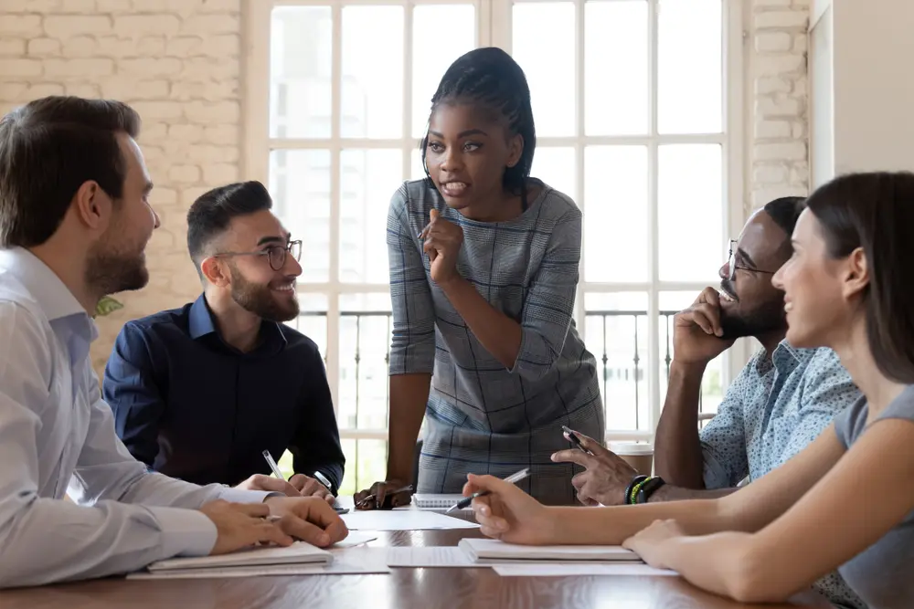 Female black executive leader talking to happy diverse employees group at corporate office briefing, multiracial coworkers listening to african woman boss explain new strategy plan at team meeting