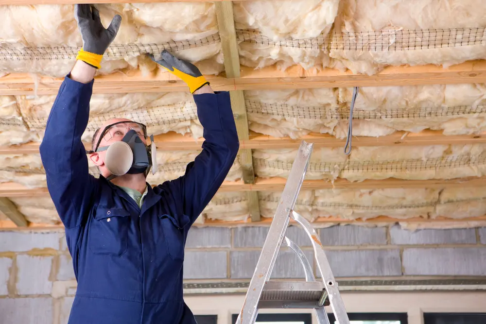 A man wearing headgear with protective mask standing on a ladder and installing ceiling insulation in an attic.