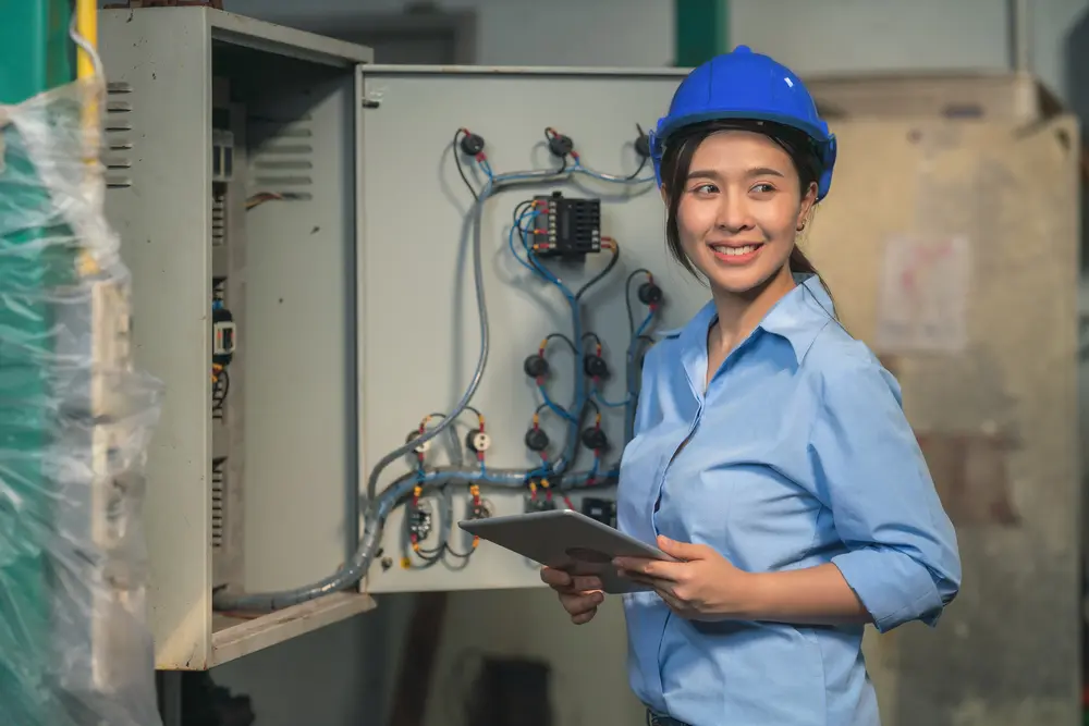 asian female industrial electrical engineer with safety hardhat and tablet on hand working in factory checking and maintenance electric control switchboard