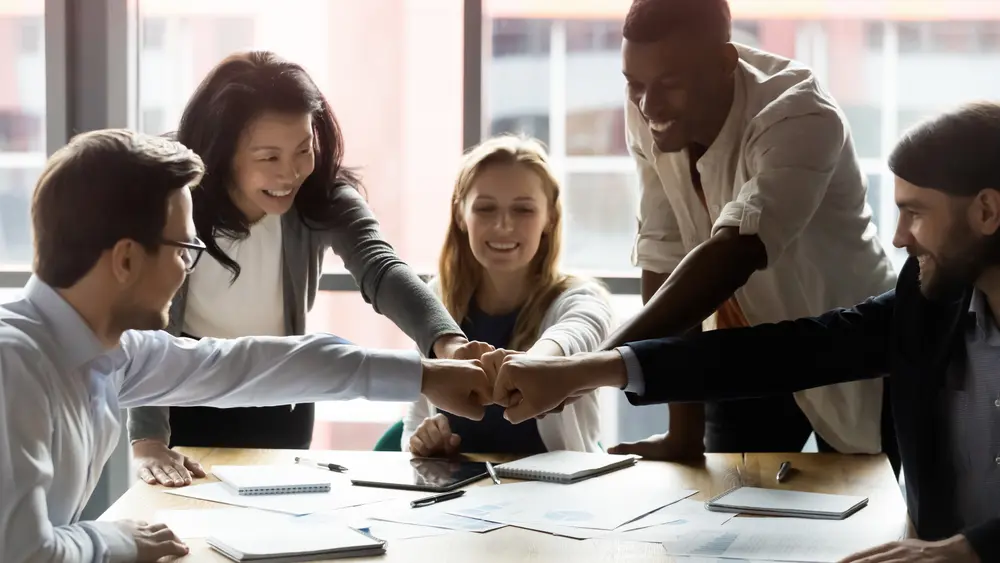 Happy motivated young and older generation multiracial business people joining fists, showing support and team spirit together, celebrating corporate achievement or success at meeting in office.