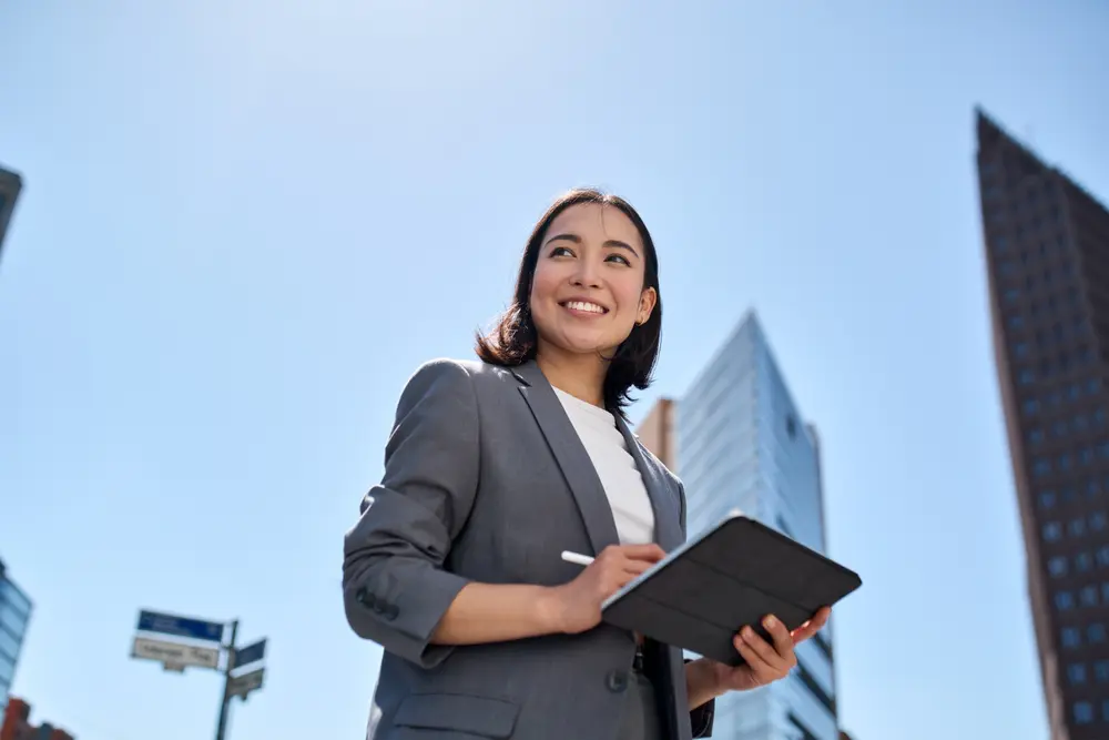 Smiling young Asian business woman leader entrepreneur, professional manager holding digital tablet computer using software applications standing on the street in big city on sky background.