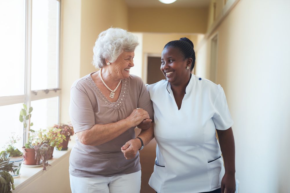 Portrait of smiling home caregiver and senior woman walking together through a corridor. Healthcare worker taking care of elderly woman.