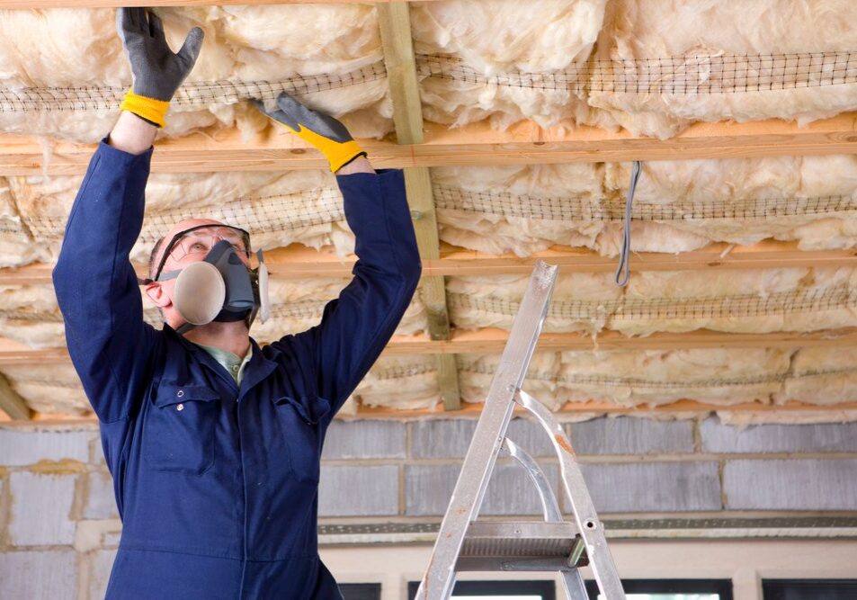 A man wearing headgear with protective mask standing on a ladder and installing ceiling insulation in an attic.