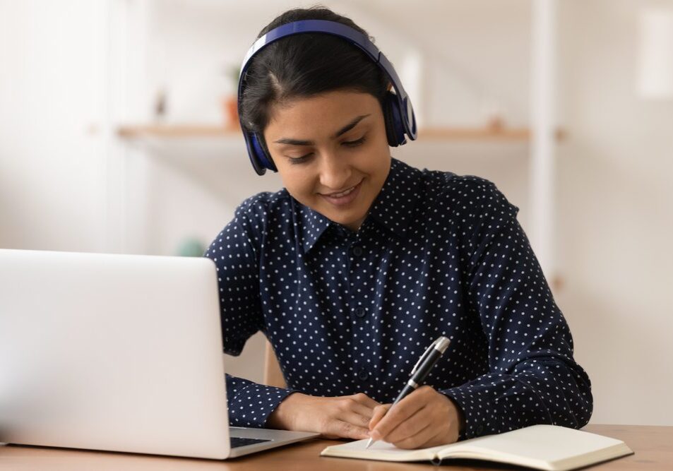 Remote student. Focused young indian lady wearing headphones practice in distant learning of english language write test before pc screen. Hindu woman in earphones listen to webinar online take notes